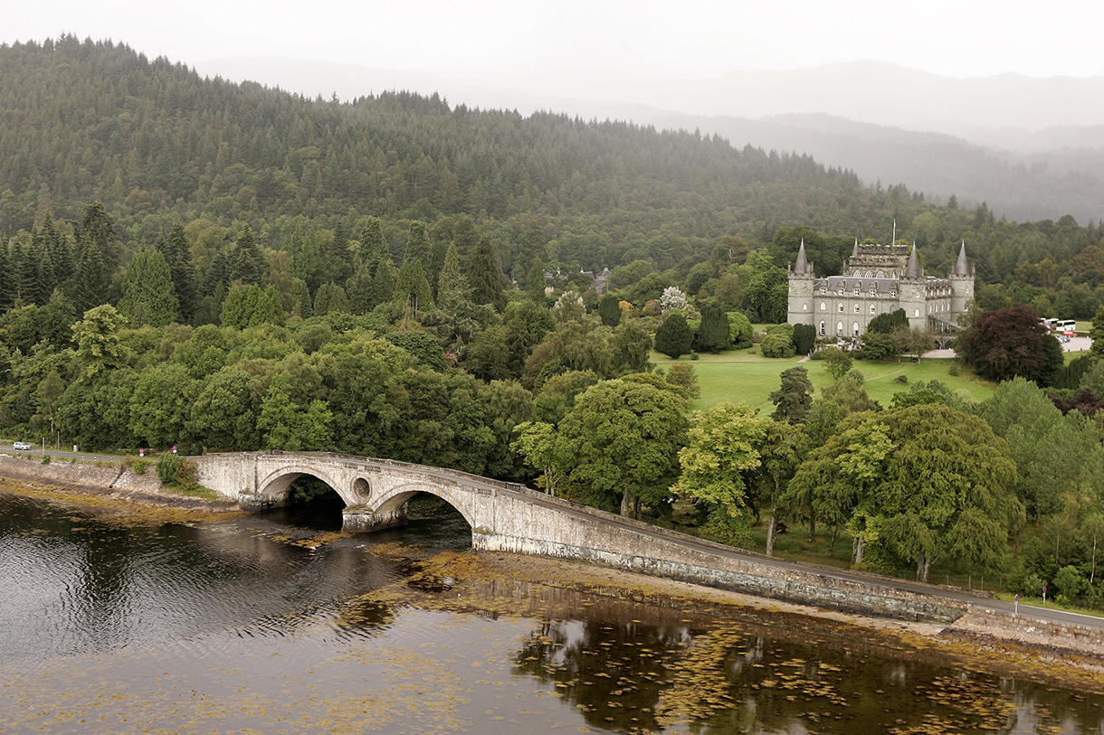 Bird’s eye view of Inveraray Castle situated in the midst of many beautiful, lush, green trees, fields and mountains. The castle is surrounded by a moat with a stone bridge.